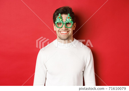 Concept of christmas, winter holidays and celebration. Close-up of handsome man in white sweater and party glasses, enjoying new year, standing over red background Concept of christmas, winter holidays and celebration. Close-up of handsome man in white sweater and party glasses, enjoying new year, standing over red background 71610079