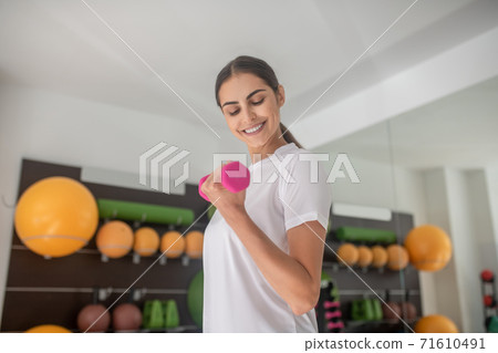 Smiling dark-haired female lifting dumbbell, looking at her biceps 71610491