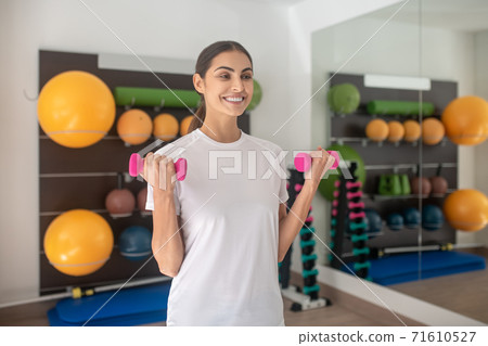 Smiling dark-haired female lifting dumbbells in the gym 71610527