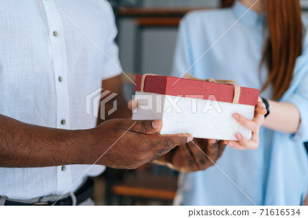 Close-up view of woman giving gift box to colleague at office. Close-up view of woman giving gift box to colleague at office. 71616534
