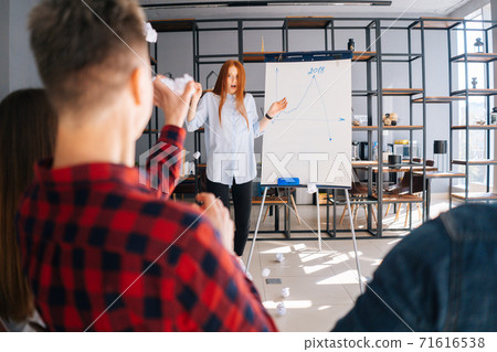 Redhead business woman finishing report on board. Dissatisfied colleagues throw crumpled paper balls Redhead business woman finishing report on board. Dissatisfied colleagues throw crumpled paper balls 71616538