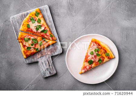 Hot pizza slices with mozzarella cheese, ham, tomato and parsley on wooden cutting board and plate, stone concrete background, top view Hot pizza slices with mozzarella cheese, ham, tomato and parsley on wooden cutting board and plate, stone concrete background, top view 71620944