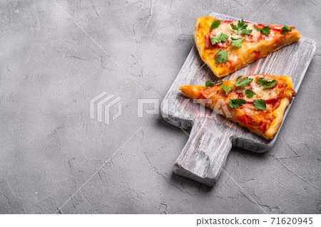 Hot pizza slices with mozzarella cheese, ham, tomato and parsley on wooden cutting board, stone concrete background, angle view copy space for text Hot pizza slices with mozzarella cheese, ham, tomato and parsley on wooden cutting board, stone concrete background, angle view copy space for text 71620945