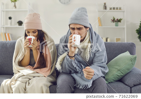 Sick young couple wrapped in plaids sitting on sofa, drinking hot tea and trying to warm up Sick young couple wrapped in plaids sitting on sofa, drinking hot tea and trying to warm up 71622069