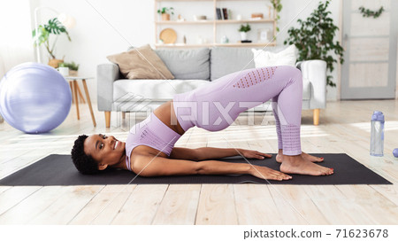 Pretty black lady doing yoga on sports mat, standing in half bridge pose at home 71623678