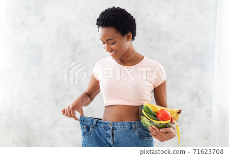 Happy African American woman in old oversized jeans holding bowl with fruits and vegetables on grey background Happy African American woman in old oversized jeans holding bowl with fruits and vegetables on grey background 71623708