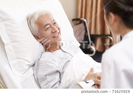 asian old man lying in bed talking to young female doctor in hospital ward 71625929