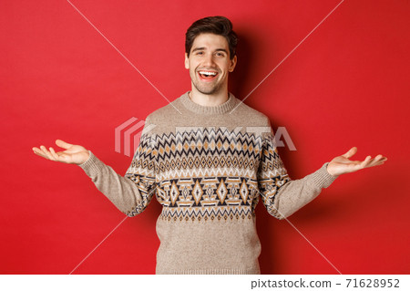 Portrait of happy good-looking man celebrating new year holidays, wearing christmas sweater, spread hands sideways and smiling, holding something on copy space, standing over red background Portrait of happy good-looking man celebrating new year holidays, wearing christmas sweater, spread hands sideways and smiling, holding something on copy space, standing over red background 71628952