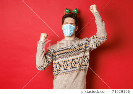 Concept of coronavirus, winter holidays and social distancing. Image of excited and happy man in medical mask and christmas clothing, raising hands up and celebrating new year, red background Concept of coronavirus, winter holidays and social distancing. Image of excited and happy man in medical mask and christmas clothing, raising hands up and celebrating new year, red background 71629106