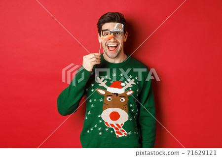 Portrait of amused handsome man in christmas sweater, holding funny party mask and smiling, celebrating winter holidays, standing over red background Portrait of amused handsome man in christmas sweater, holding funny party mask and smiling, celebrating winter holidays, standing over red background 71629121
