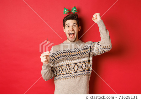 Cheerful guy with a sparkler rejoicing and wishing happy new year, raising hand up and shouting for joy, standing over red background 71629131