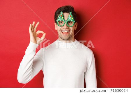 Happy caucasian guy wishing merry christmas, celebrating winter holidays, showing okay sign and smiling pleased, standing over red background Happy caucasian guy wishing merry christmas, celebrating winter holidays, showing okay sign and smiling pleased, standing over red background 71630024