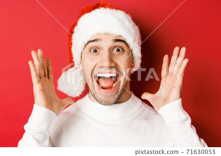 Close-up of happy young man in santa hat, making big christmas announcement, smiling amazed, standing over red background 71630055