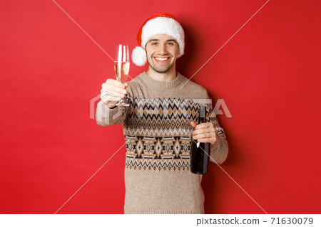 Concept of winter holidays, new year and celebration. Portrait of handsome man in santa hat and sweater, holding champagne, raising glass and saying cheers on christmas party Concept of winter holidays, new year and celebration. Portrait of handsome man in santa hat and sweater, holding champagne, raising glass and saying cheers on christmas party 71630079