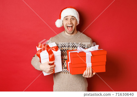 Concept of winter holidays, new year and celebration. Image of amazed and happy man in sweater and santa hat, holding christmas gifts and shouting for joy, red background Concept of winter holidays, new year and celebration. Image of amazed and happy man in sweater and santa hat, holding christmas gifts and shouting for joy, red background 71630640