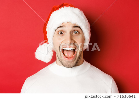Concept of winter holidays, christmas and celebration. Close-up of surprised handsome man in santa hat, hear amazing new year promo offer, standing against red background Concept of winter holidays, christmas and celebration. Close-up of surprised handsome man in santa hat, hear amazing new year promo offer, standing against red background 71630641