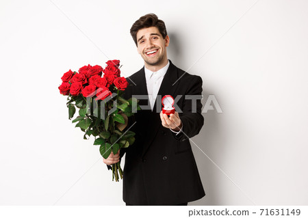 Handsome smiling man in black suit, holding roses and engagement ring, making a proposal to marry him, standing against white background Handsome smiling man in black suit, holding roses and engagement ring, making a proposal to marry him, standing against white background 71631149