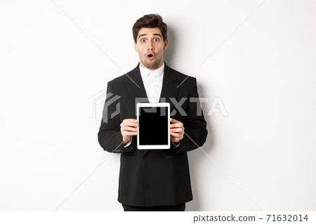 Image of surprised handsome man in black suit, showing digital tablet screen and looking amazed, standing against white background 71632014