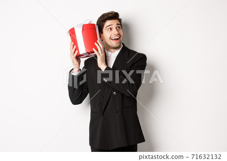 Concept of christmas holidays, celebration and lifestyle. Image of attractive man in black suit, shaking present to guess what inside, enjoying new year, standing against white background Concept of christmas holidays, celebration and lifestyle. Image of attractive man in black suit, shaking present to guess what inside, enjoying new year, standing against white background 71632132