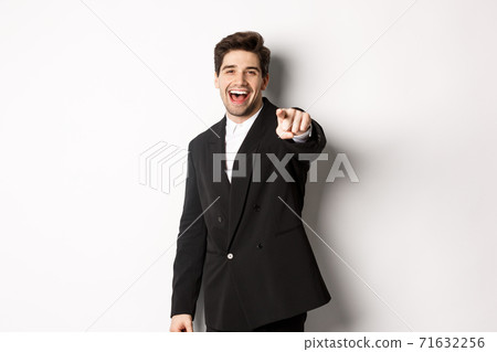 Concept of new year party, celebration and lifestyle. Portrait of handsome stylish man in black suit, smiling and pointing finger at camera, standing over white background Concept of new year party, celebration and lifestyle. Portrait of handsome stylish man in black suit, smiling and pointing finger at camera, standing over white background 71632256