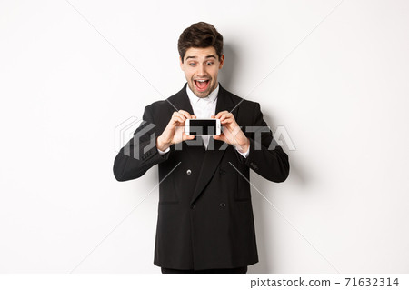 Portrait of excited handsome man showing smartphone screen, looking amazed at display with advertisement, standing over white background in black suit 71632314