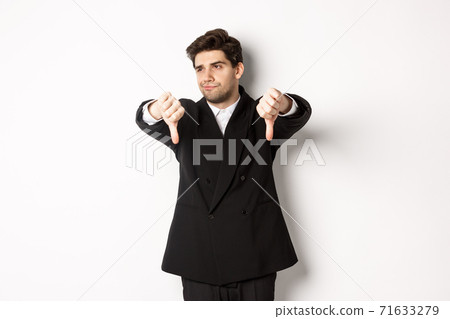 Skeptical and displeased man in black suit, looking away reluctant, showing thumbs-down, dislike something, standing over white background Skeptical and displeased man in black suit, looking away reluctant, showing thumbs-down, dislike something, standing over white background 71633279