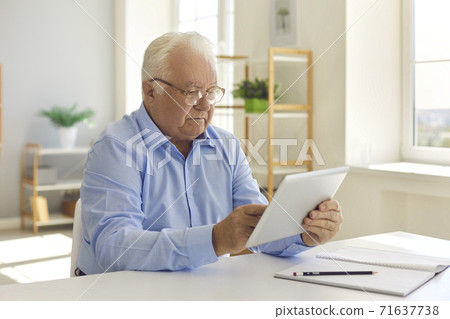 Senior man with glasses sitting at a table at home using a tablet reads information on the Internet. Senior man with glasses sitting at a table at home using a tablet reads information on the Internet. 71637738