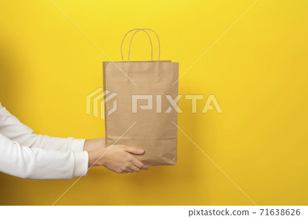 Female hands hold large gift bag made of brown craft paper on yellow background. Female hands hold large gift bag made of brown craft paper on yellow background. 71638626
