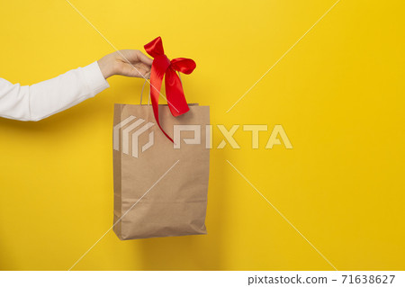 Female hand hold large gift bag made of brown craft paper with a red bow. Female hand hold large gift bag made of brown craft paper with a red bow. 71638627