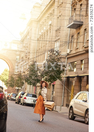 Full length of a stylish happy woman wearing grey coat, hat and high heel shoes standing with shopping bags on city street and smiling at camera 71639274