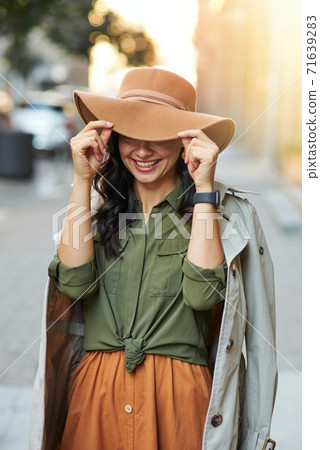 Playful beauty. Young happy caucasian woman adjusting hat and smiling while standing on the city street, enjoying walking on warm autumn day Playful beauty. Young happy caucasian woman adjusting hat and smiling while standing on the city street, enjoying walking on warm autumn day 71639283