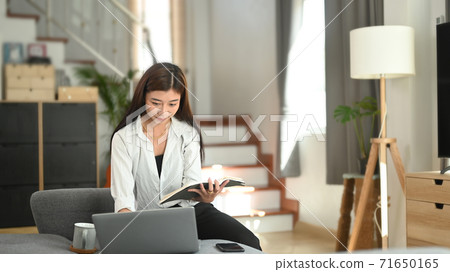 A young female in casual clothes is reading a book while using tablet on couch at home. A young female in casual clothes is reading a book while using tablet on couch at home. 71650165