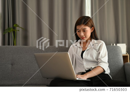 A beautiful cheerful is girl using silver laptop while sitting on sofa in living room at home. A beautiful cheerful is girl using silver laptop while sitting on sofa in living room at home. 71650303