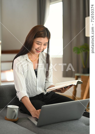 A happy woman reading a book while using laptop on sofa in her living room. A happy woman reading a book while using laptop on sofa in her living room. 71650374