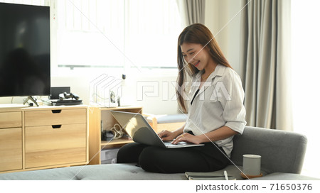 A happy smiling woman sitting on sofa and using laptop at living room at home. A happy smiling woman sitting on sofa and using laptop at living room at home. 71650376