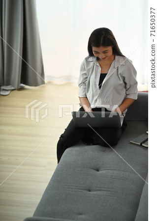 A beautiful asian woman enjoying time and sitting on modern sofa relaxing with laptop in her living room. A beautiful asian woman enjoying time and sitting on modern sofa relaxing with laptop in her living room. 71650377