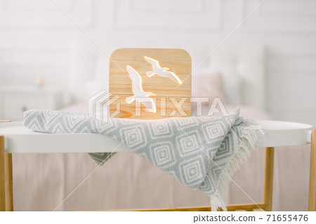 Front close up view of stylish wooden night lamp with birds picture, on gray blanket at cozy light bedroom interior. Wooden decorations at home interior Front close up view of stylish wooden night lamp with birds picture, on gray blanket at cozy light bedroom interior. Wooden decorations at home interior 71655476