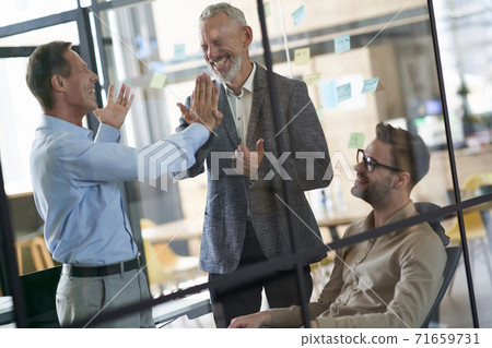 Two excited mature businessmen giving high five to each other while having a meeting with colleagues in the modern office 71659731