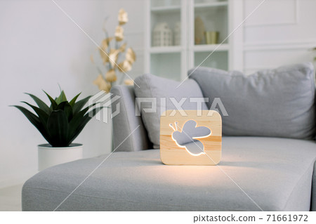 Home decor and lamps. Wooden night lamp with bee picture, lying on gray modern sofa, at stylish light home living room interior 71661972