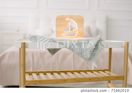 Wooden lapms and decor details at home interior. Stylish wooden hand made lamp with ship cut out picture, on little coffee table, standing in light home bedroom 71662019