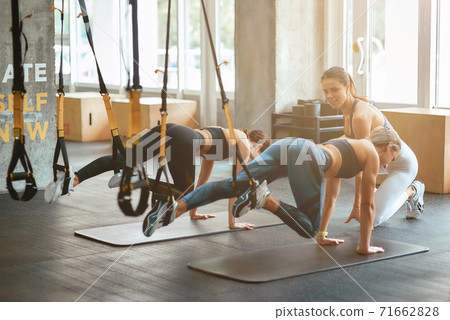Two young athletic girls exercising with trx fitness straps at industrial gym with assistance of personal trainer 71662828