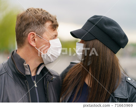 Hugging heterosexual couple in love of young handsome man and pretty woman in rainy spring day. They look at each other. Wearing of protective face masks during coronavirus pandemic. Hugging heterosexual couple in love of young handsome man and pretty woman in rainy spring day. They look at each other. Wearing of protective face masks during coronavirus pandemic. 71663969