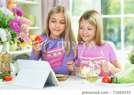 Two girls in pink aprons preparing salad on kitchen table with tablet Two girls in pink aprons preparing salad on kitchen table with tablet 71669293