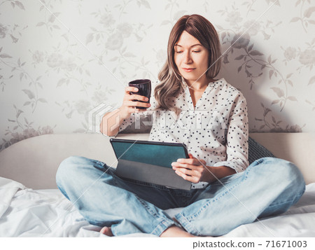 Woman sits on bed with tablet PC and cup of hot coffee. She watches online TV series. Online video call or conference, distance learning, remote education. Self isolation during quarantine. 71671003