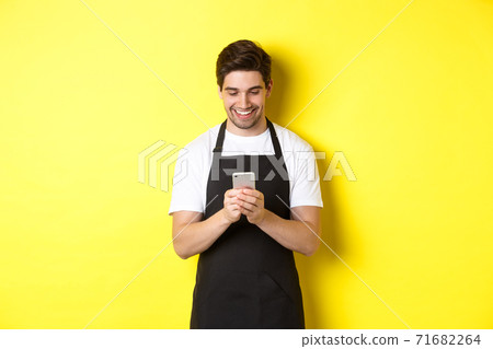Waiter in black apron reading message on mobile phone, smiling happy, standing over yellow background Waiter in black apron reading message on mobile phone, smiling happy, standing over yellow background 71682264