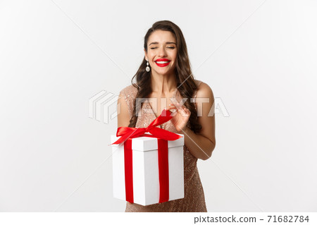 Merry Christmas. Charming woman in elegant dress open gift box and smiling delighted, unwrapping a present, standing over white background Merry Christmas. Charming woman in elegant dress open gift box and smiling delighted, unwrapping a present, standing over white background 71682784