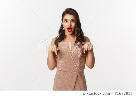 Amazed beautiful woman in glamour dress celebrating christmas party, pointing fingers down at promo, standing over white background 71682995