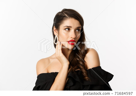 Fashion and beauty concept. Close-up of elegant woman in black dress, showing earrings and looking sensual, red lipstick and makeup on, white background 71684597