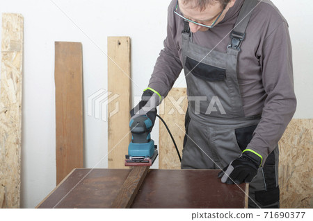 Carpenter working with  electric sheet finishing sander  on wood lath on desk, on background oriented strand board 71690377