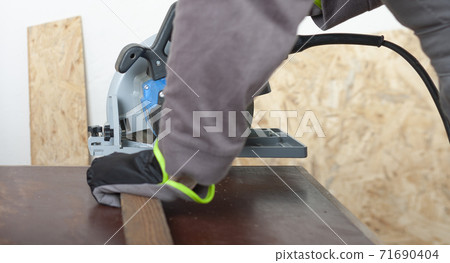 Carpenter cuts wood lath electric hand tool circular saw on desk, on background oriented strand board Carpenter cuts wood lath electric hand tool circular saw on desk, on background oriented strand board 71690404